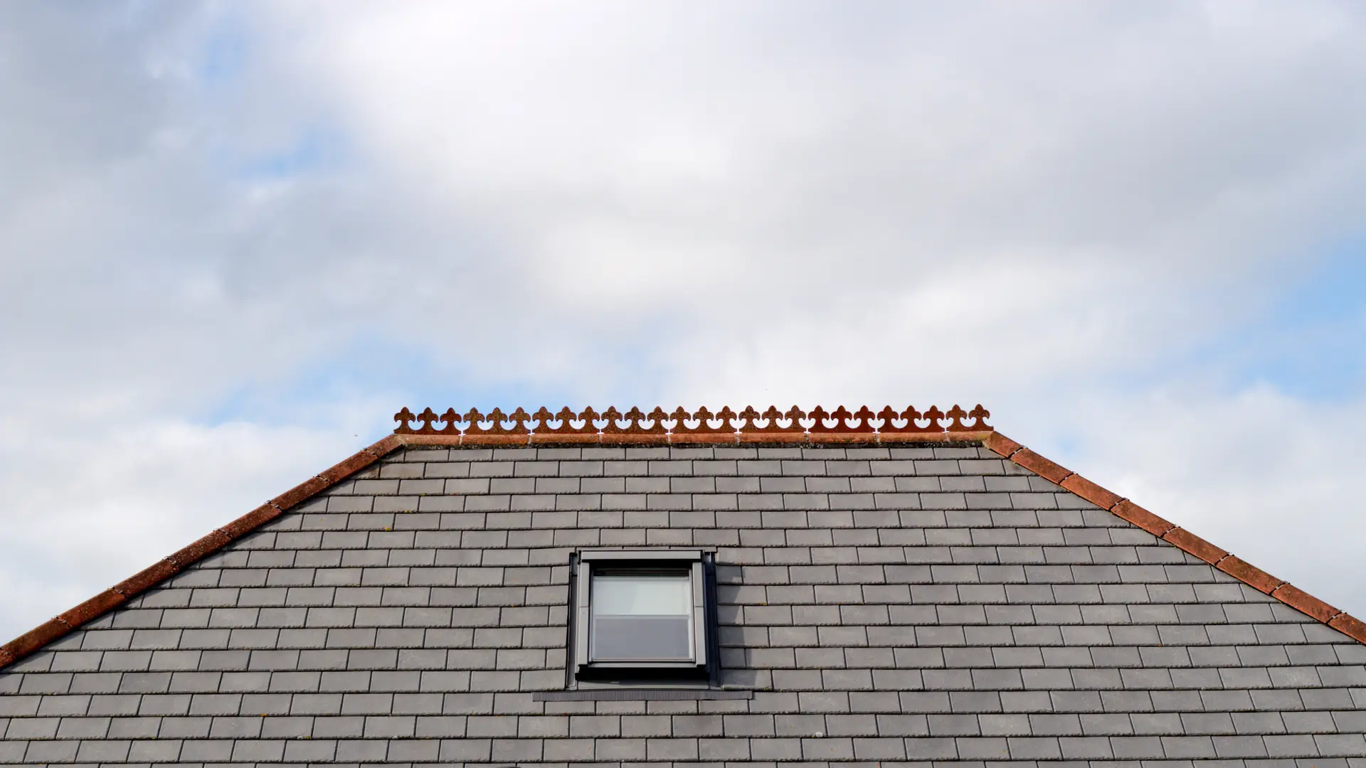 Hipped slate roof with a roof window and a clay ridge under a bright sky
